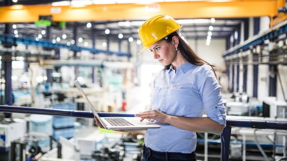 Engineer with hard hat working on laptop in modern manufacturing facility, monitoring packaging automation and production line efficiency.