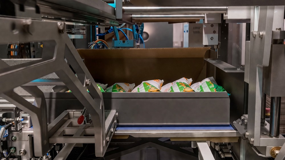 Tronrud Packaging: Close-up of an automated secondary packaging station with snack bags and an open corrugated case on a conveyor.